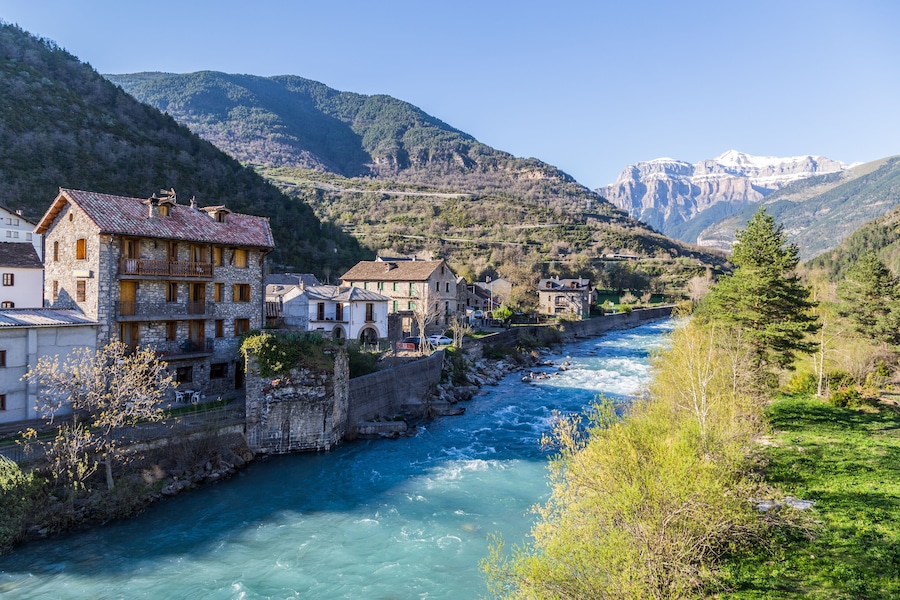 Une rivière traverse une ville où les bâtiments sont en pierre, avec des montagnes en arrière-plan.
