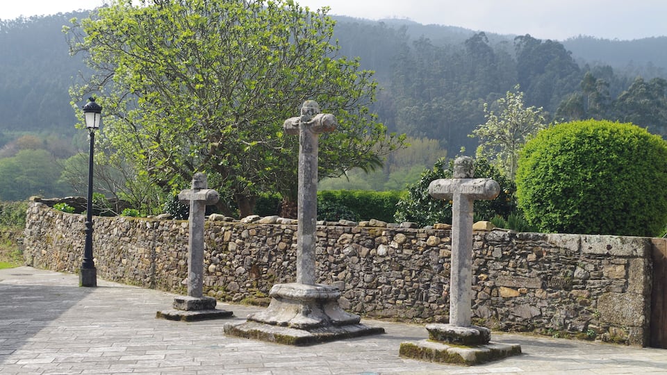 Three stone crosses on a paved path with a stone wall and a lamp post in the background.