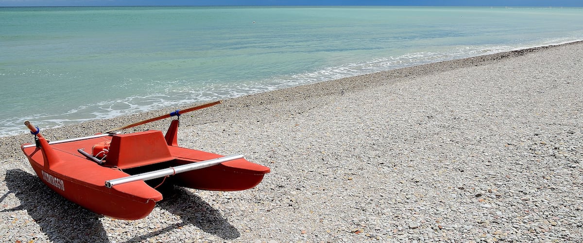 A red boat on a pebbled beach with calm turquoise waters and a clear sky.