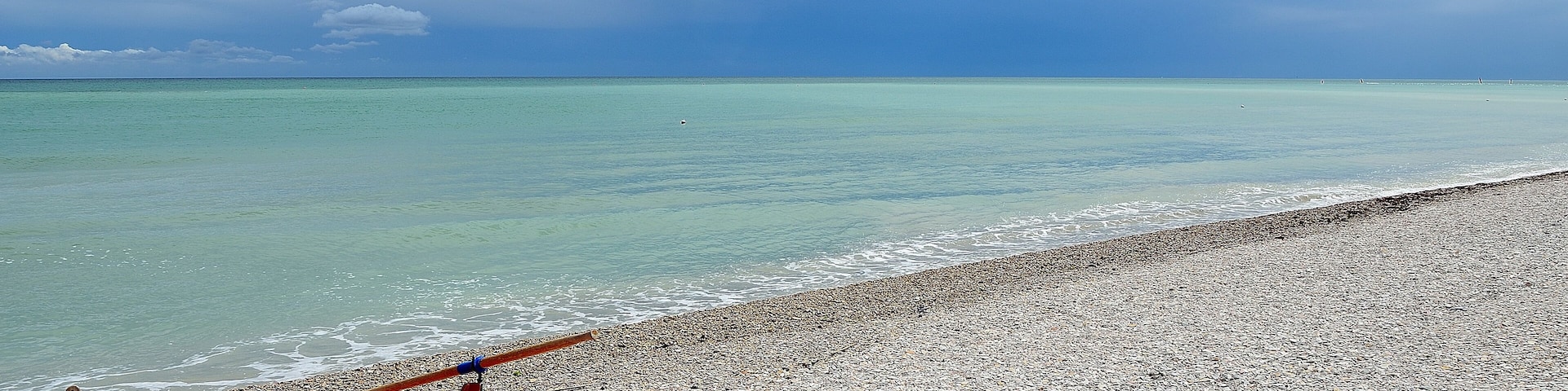 A red boat on a pebbled beach with calm turquoise waters and a clear sky.