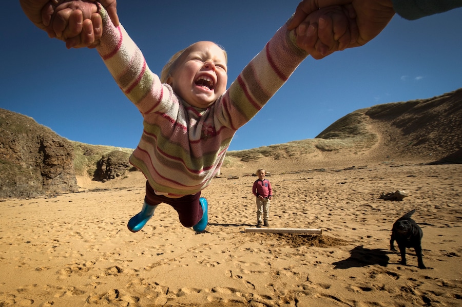 Parent holding daughter's hands, spinning her around, young son in background, low angle view, Lahinch, Clare, Ireland