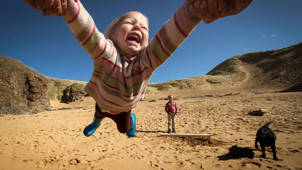 Parent holding daughter's hands, spinning her around, young son in background, low angle view, Lahinch, Clare, Ireland