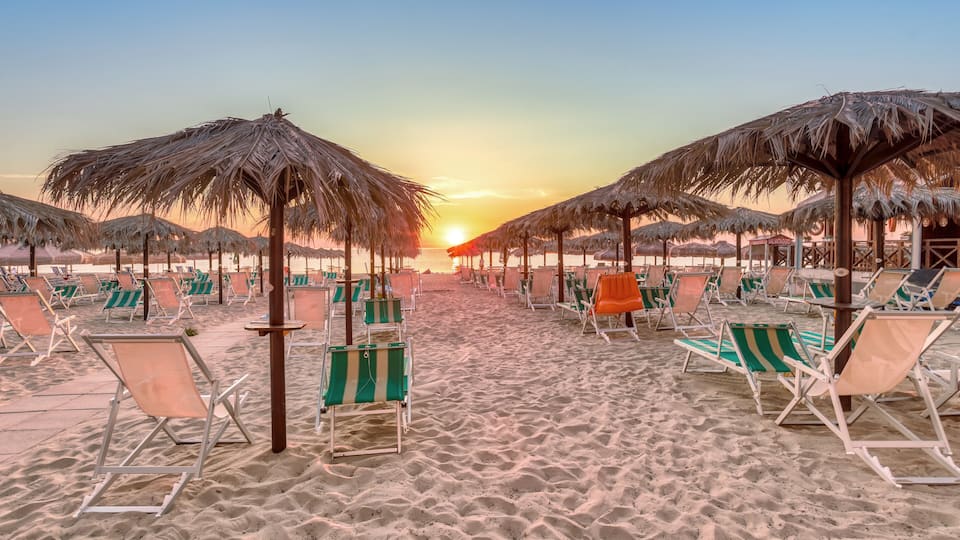A beach with rows of beach chairs and thatched umbrellas under a sunset sky.