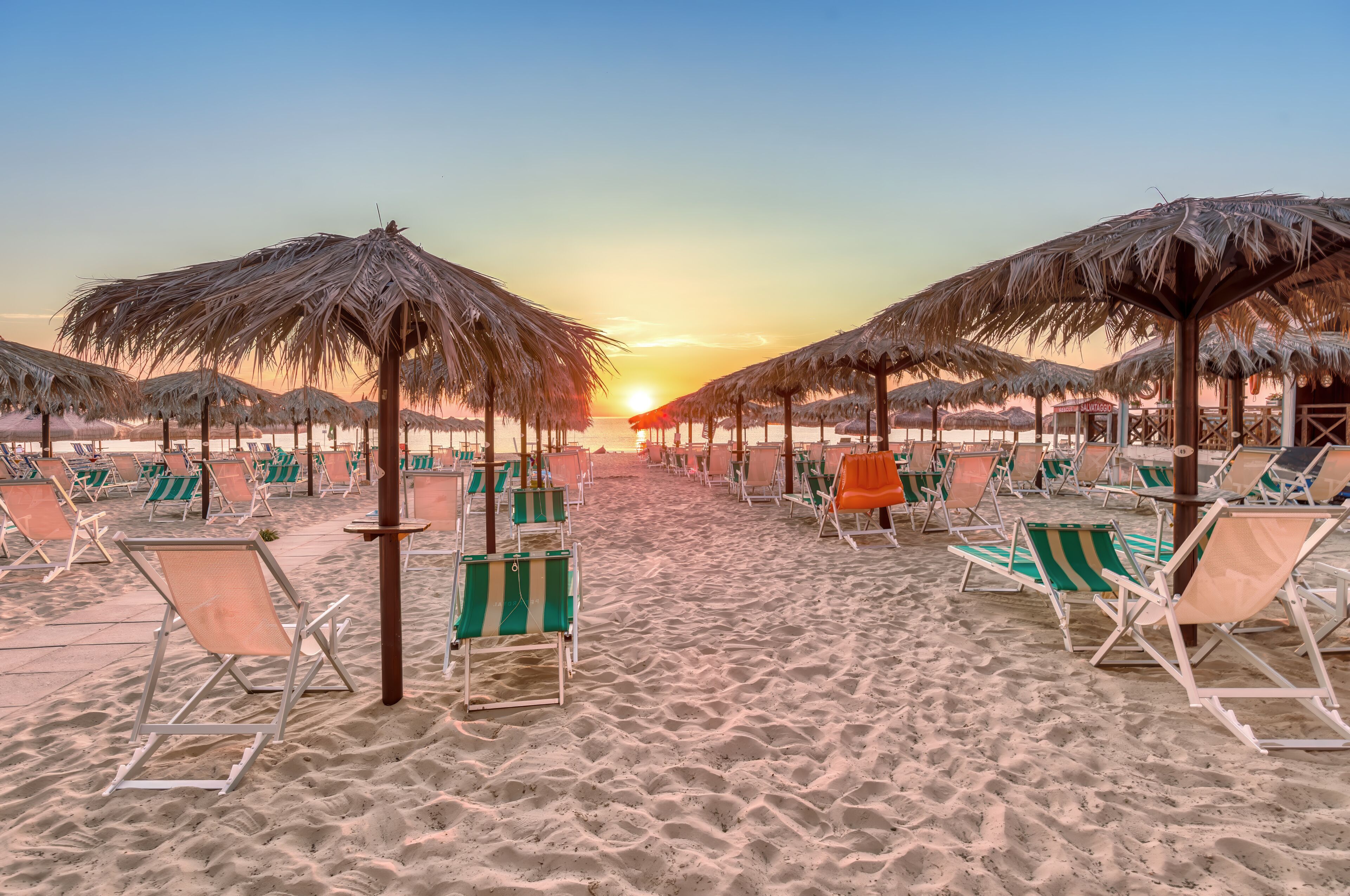 A beach with rows of beach chairs and thatched umbrellas under a sunset sky.