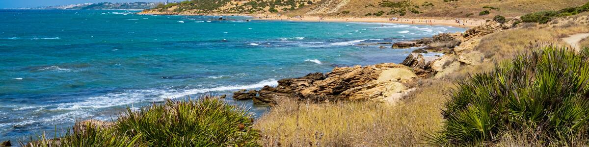 A coastal landscape with clear blue waters, rocky shores, and a hilly terrain with sparse vegetation.
