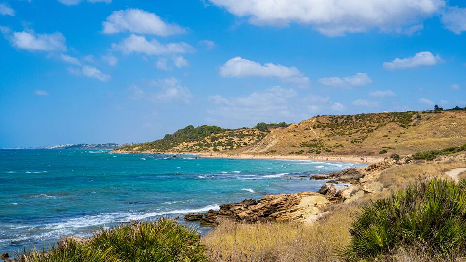 A coastal landscape with clear blue waters, rocky shores, and a hilly terrain with sparse vegetation.