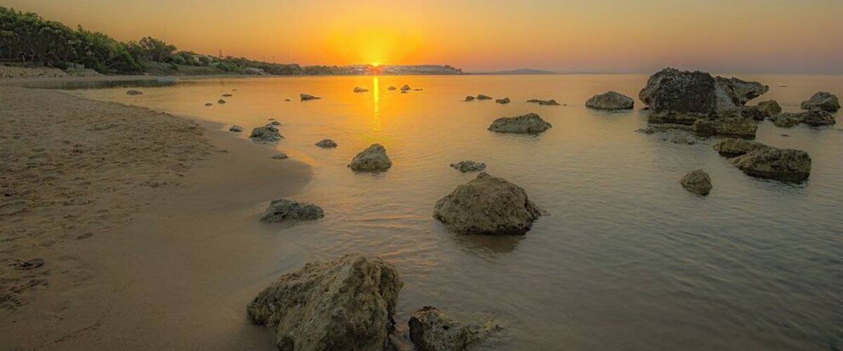 Un coucher de soleil sur une plage paisible, parsemée de rochers, avec une côte lointaine à l’horizon.