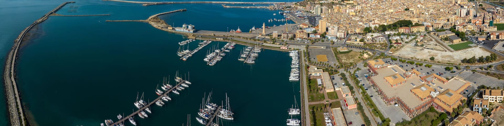 An aerial view of a coastal city with a marina filled with boats, a lighthouse, and a hilly landscape in the background.