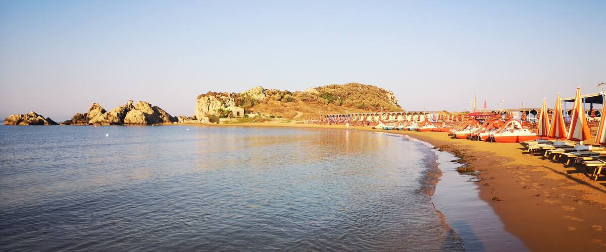A beach with calm waters, a rocky outcrop, and rows of beach umbrellas.