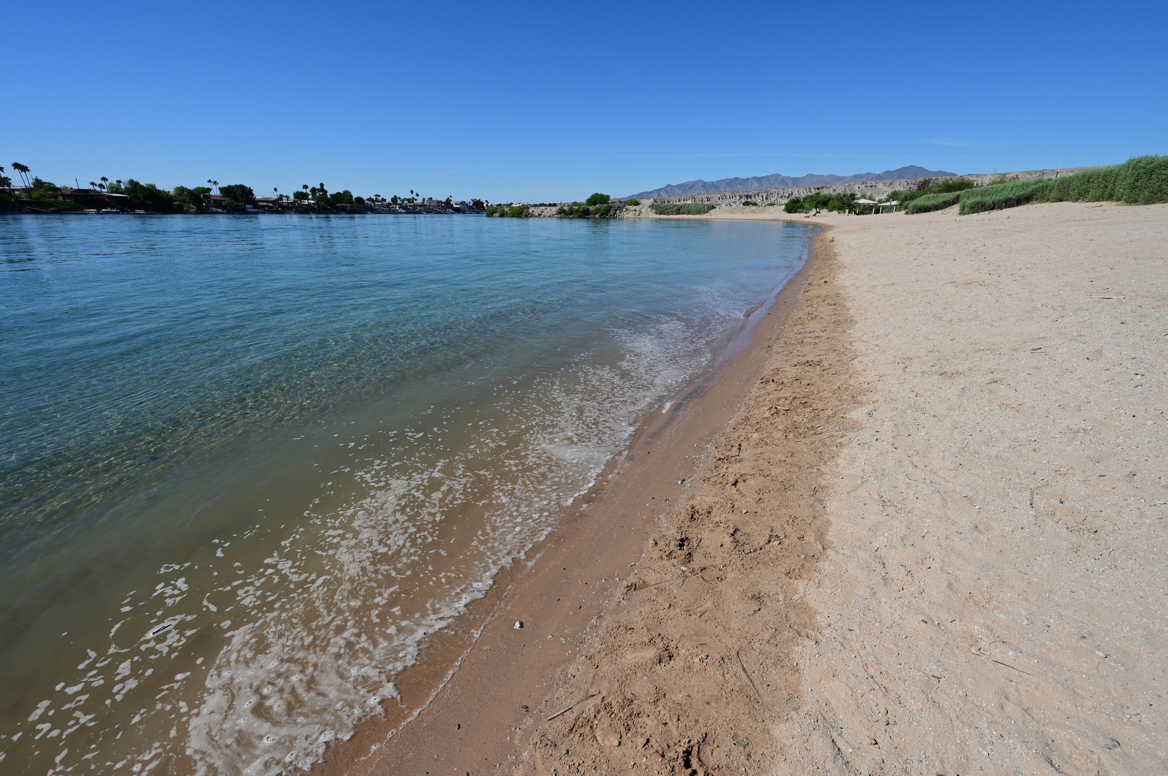 Orilla de río con aguas cristalinas, una playa arenosa y montañas a lo lejos.