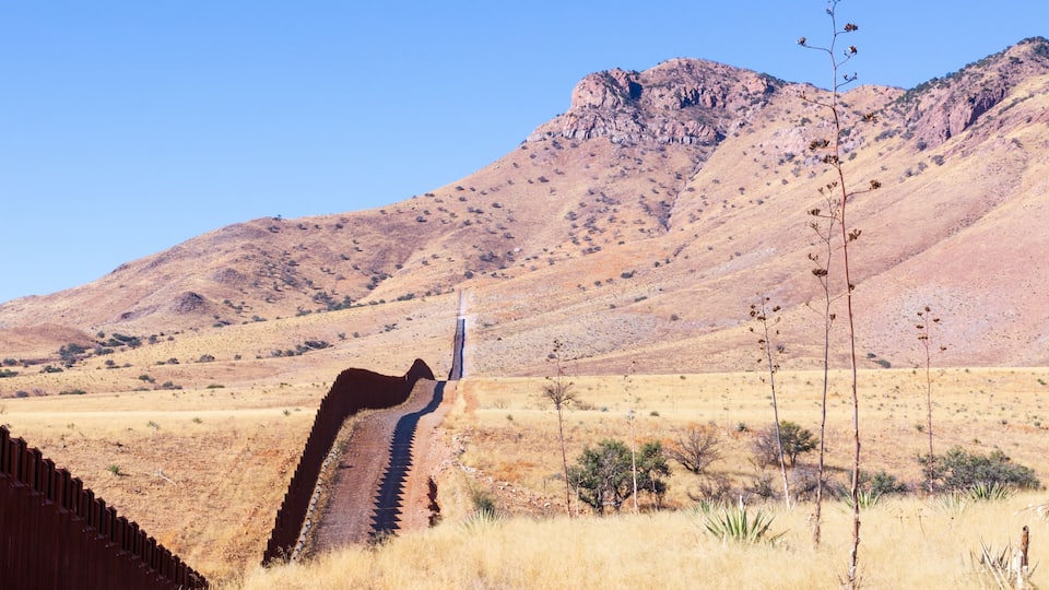 A desert landscape with a prominent mountain in the background, a fence running along a road, and sparse vegetation.
