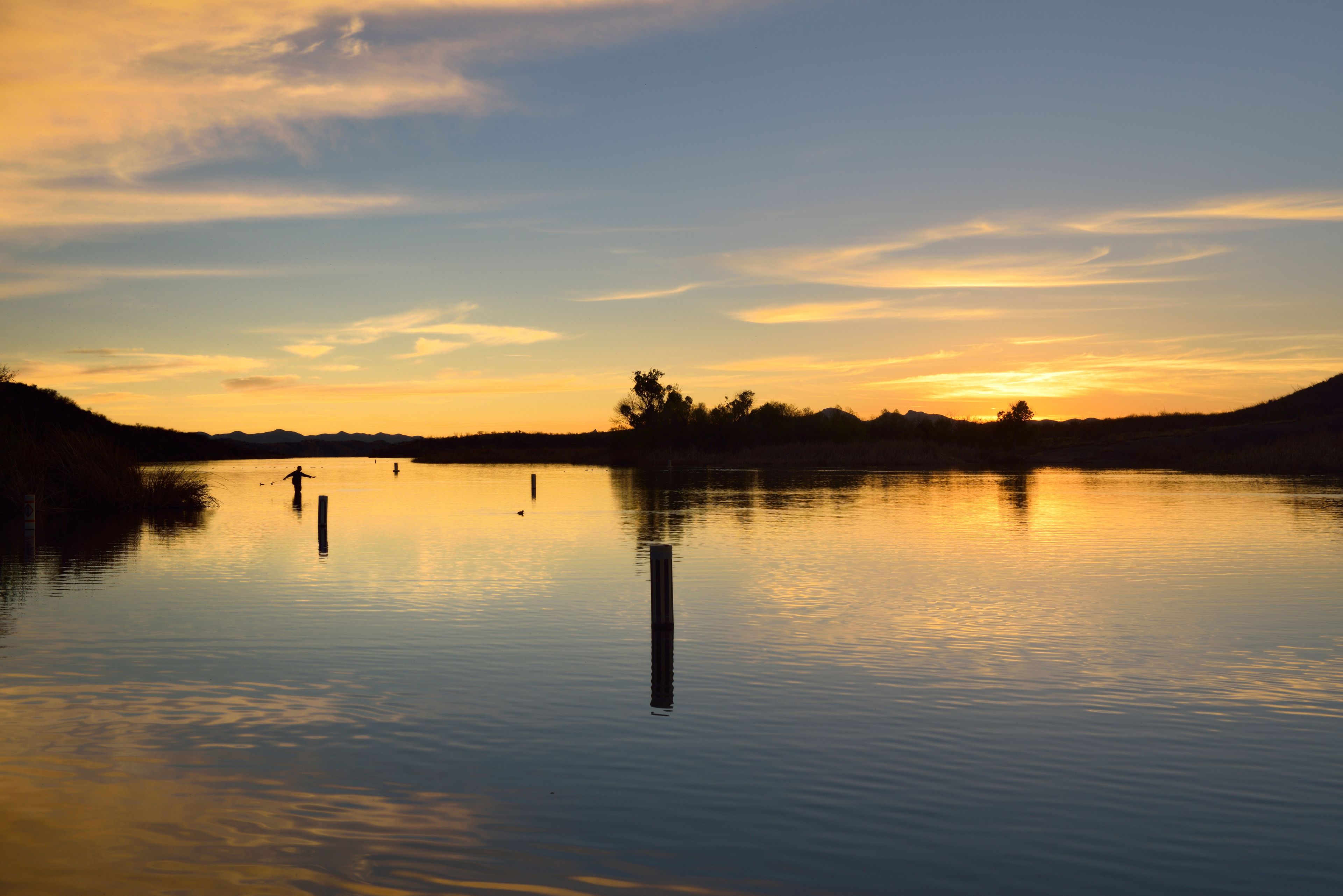 A calm lake at sunset with a silhouette of a person fishing, trees, and a clear sky.
