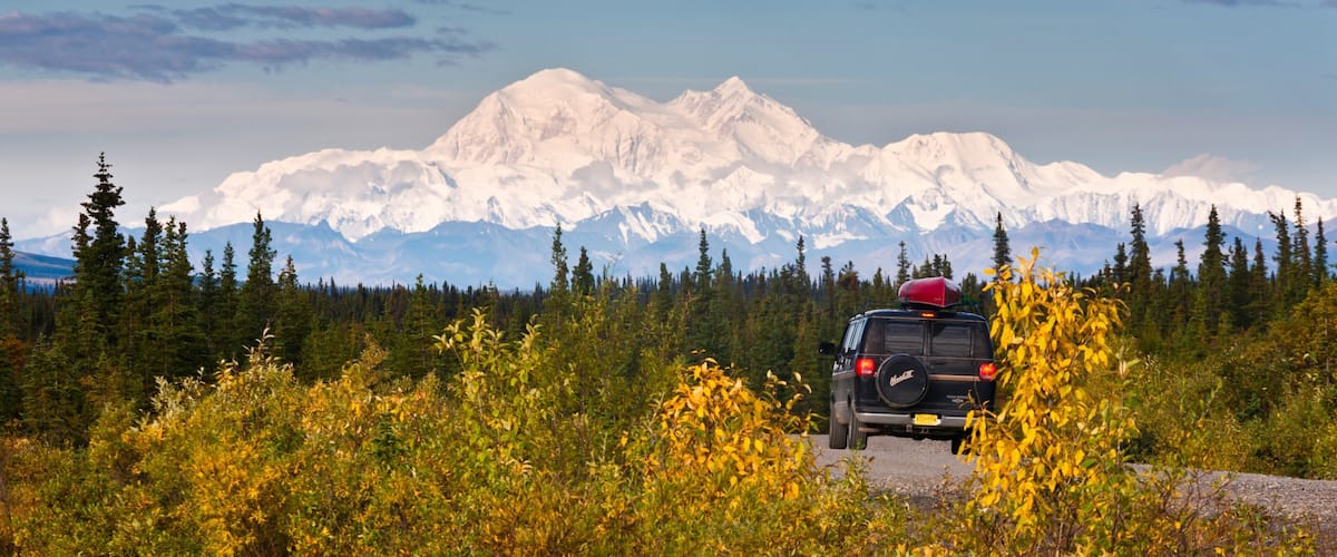 A vehicle with a red roof rack on a forested road with yellow foliage, facing snow-capped mountains.