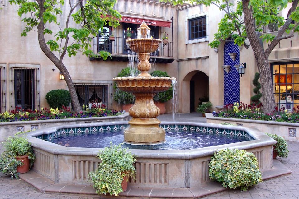 Un patio con una fuente central, rodeado de plantas en macetas y un edificio con toldo rojo.