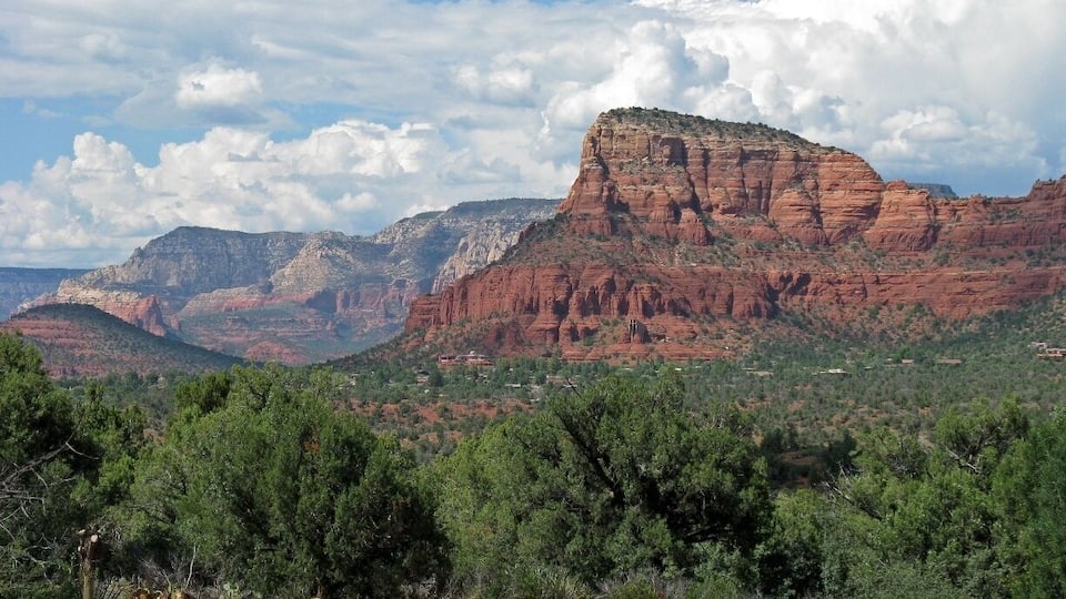 A red rock canyon with a prominent peak, surrounded by greenery and distant mountains.