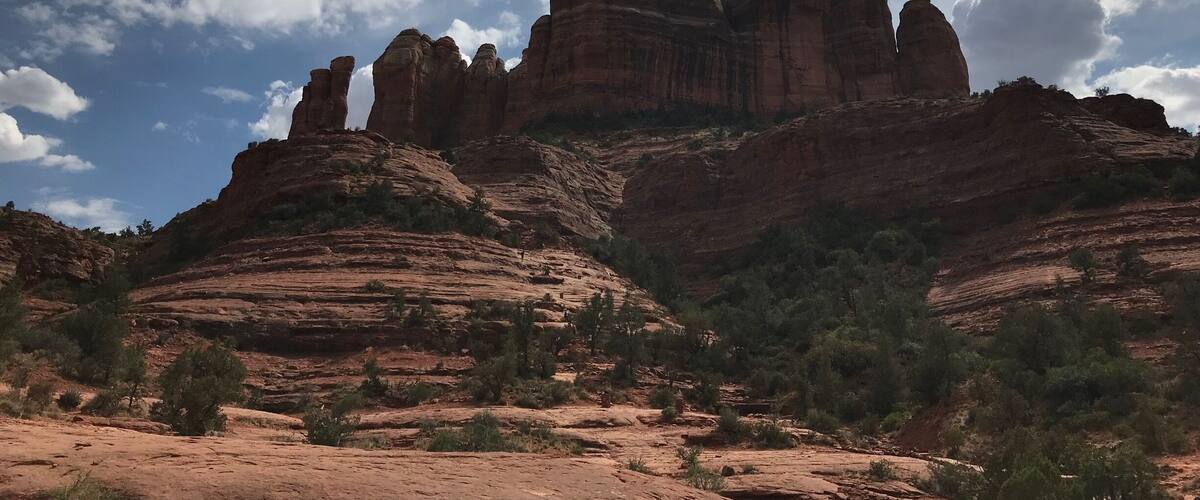 A rugged red rock formation with layered cliffs and sparse vegetation under a partly cloudy sky.