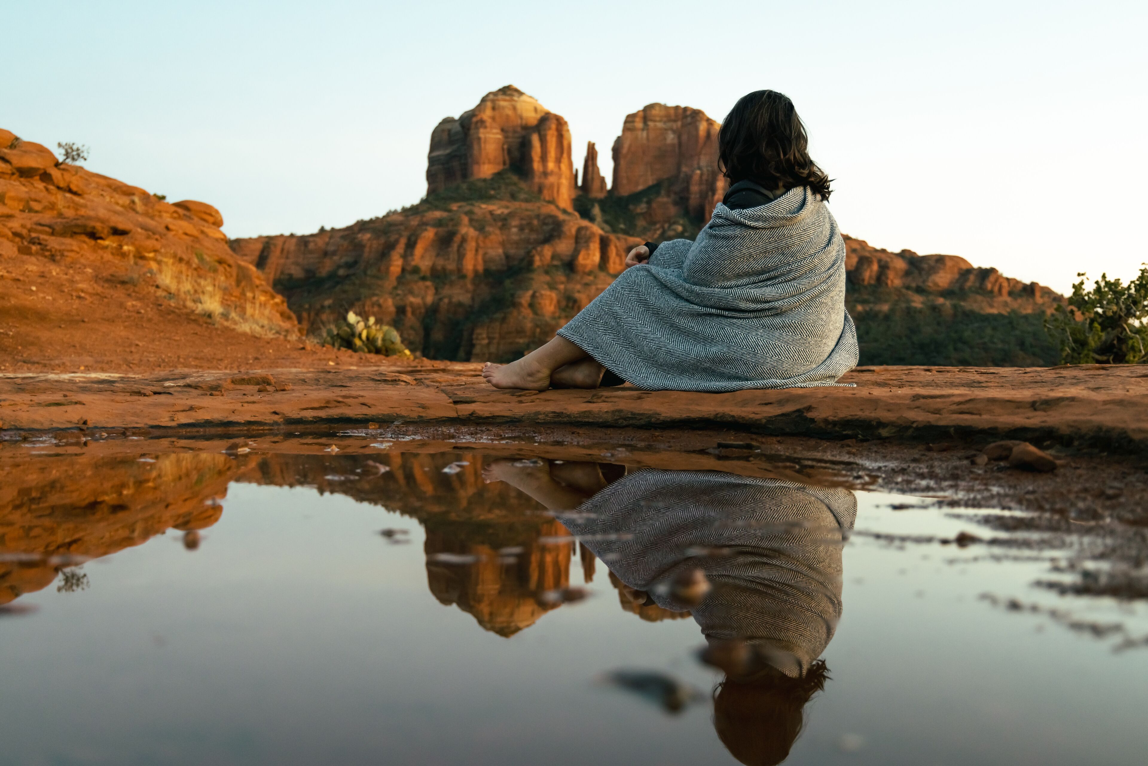 Close up view of young brunette woman 30s sitting by small pool of water on red slick rock with Cathedral Rock in Sedona Arizona USA in background.