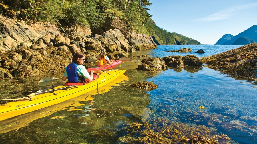 A kayaker in a yellow kayak on a rocky shoreline with clear water and a forested mountain in the background.