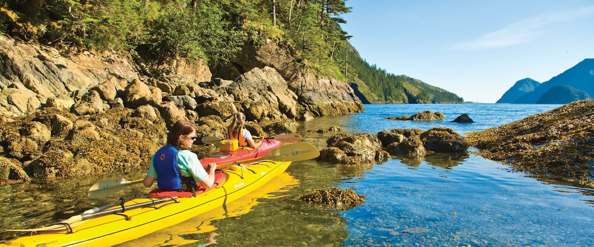 A kayaker in a yellow kayak on a rocky shoreline with clear water and a forested mountain in the background.