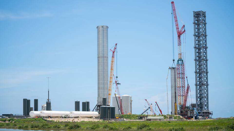 A construction site with multiple cranes, large storage tanks, and industrial buildings.