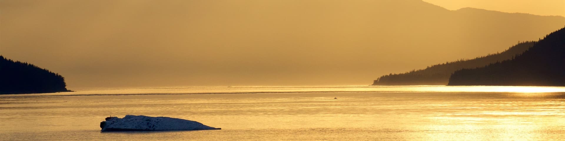 A sunset over a calm sea with a small iceberg in the foreground and mountains in the background.
