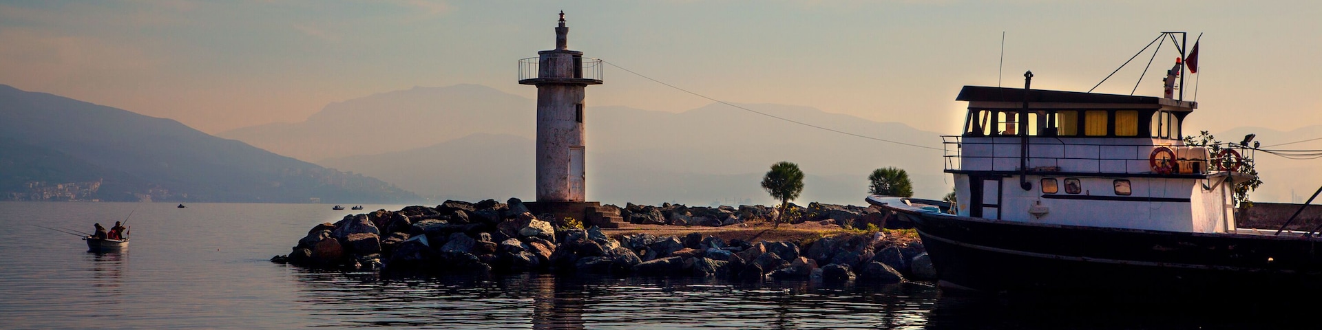 Un puerto tranquilo con un faro, un barco amarrado a la derecha y un rompeolas rocoso.