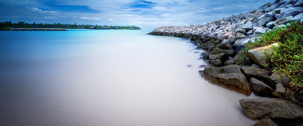 A calm beach with clear water, a rocky shoreline, and a distant tree line.