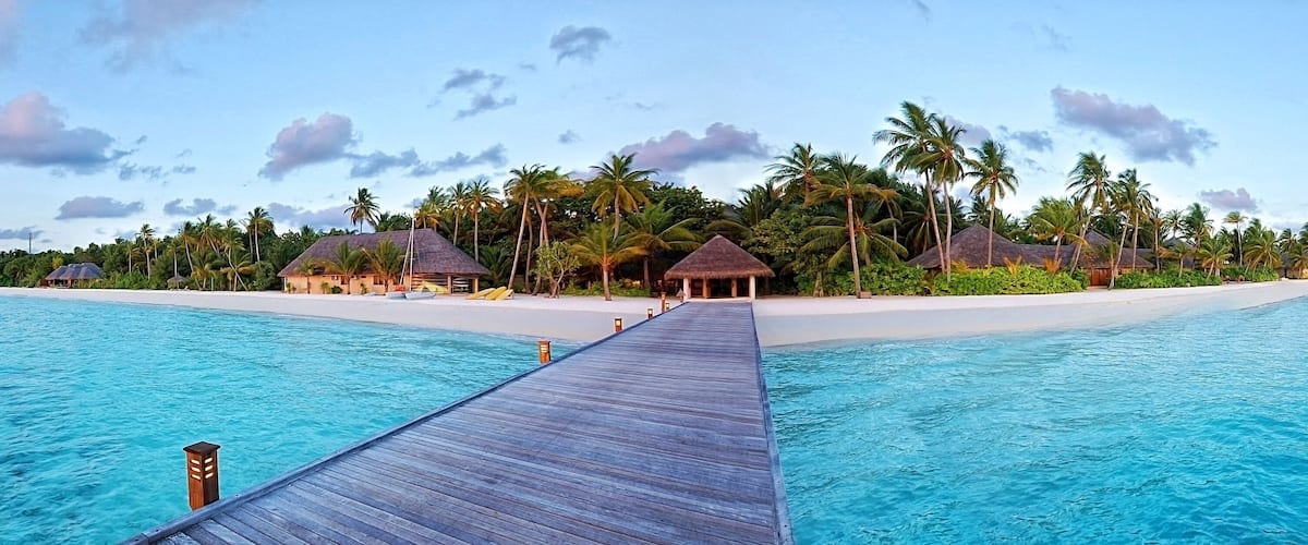 A wooden pier leading to overwater bungalows with thatched roofs, surrounded by palm trees and a clear blue sea.
