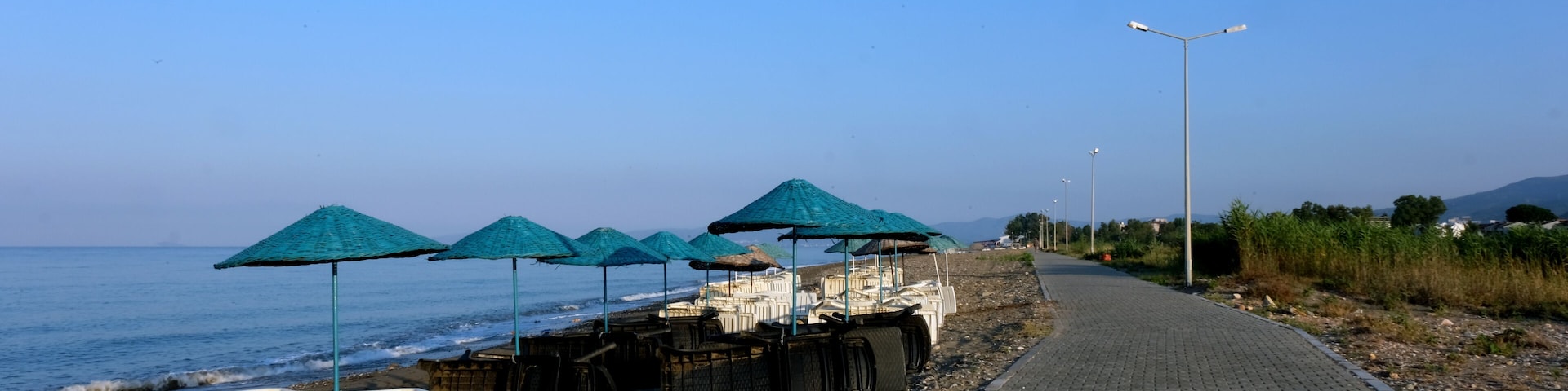 Uma praia com fileiras de guarda-sóis azuis, um calçadão pavimentado e um mar calmo.