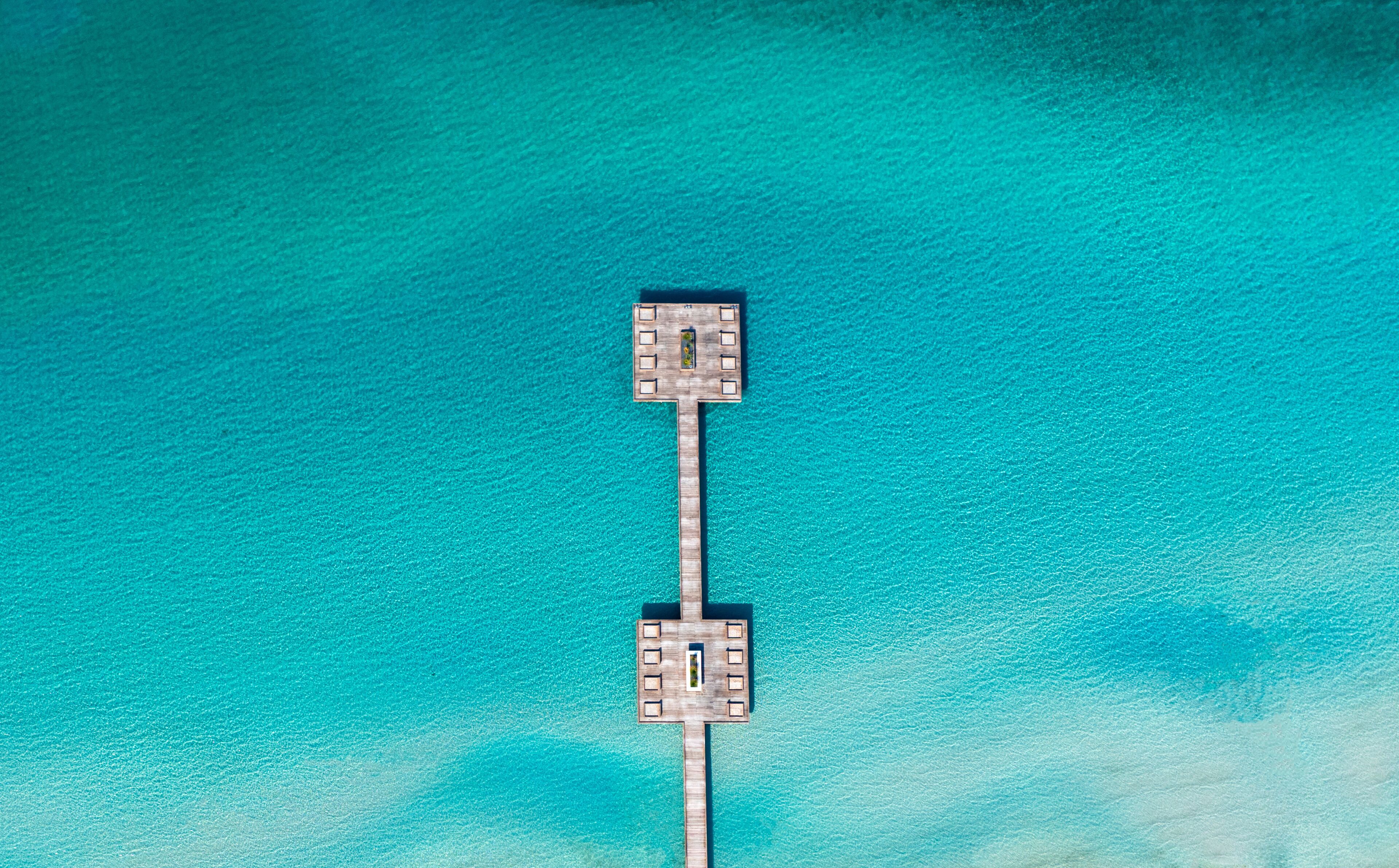 Vista aérea de un muelle de madera que se extiende sobre aguas turquesas, con un pequeño embarcadero al final.