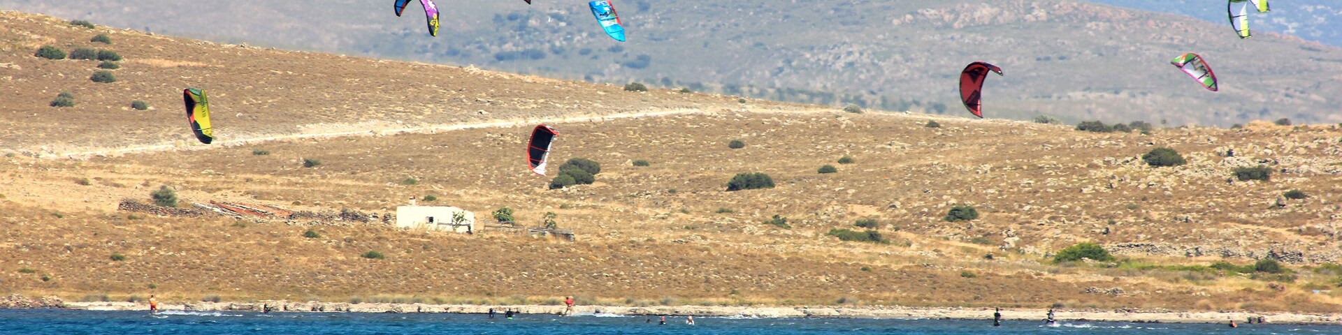 A coastal landscape with kites flying over the sea and hills in the background.