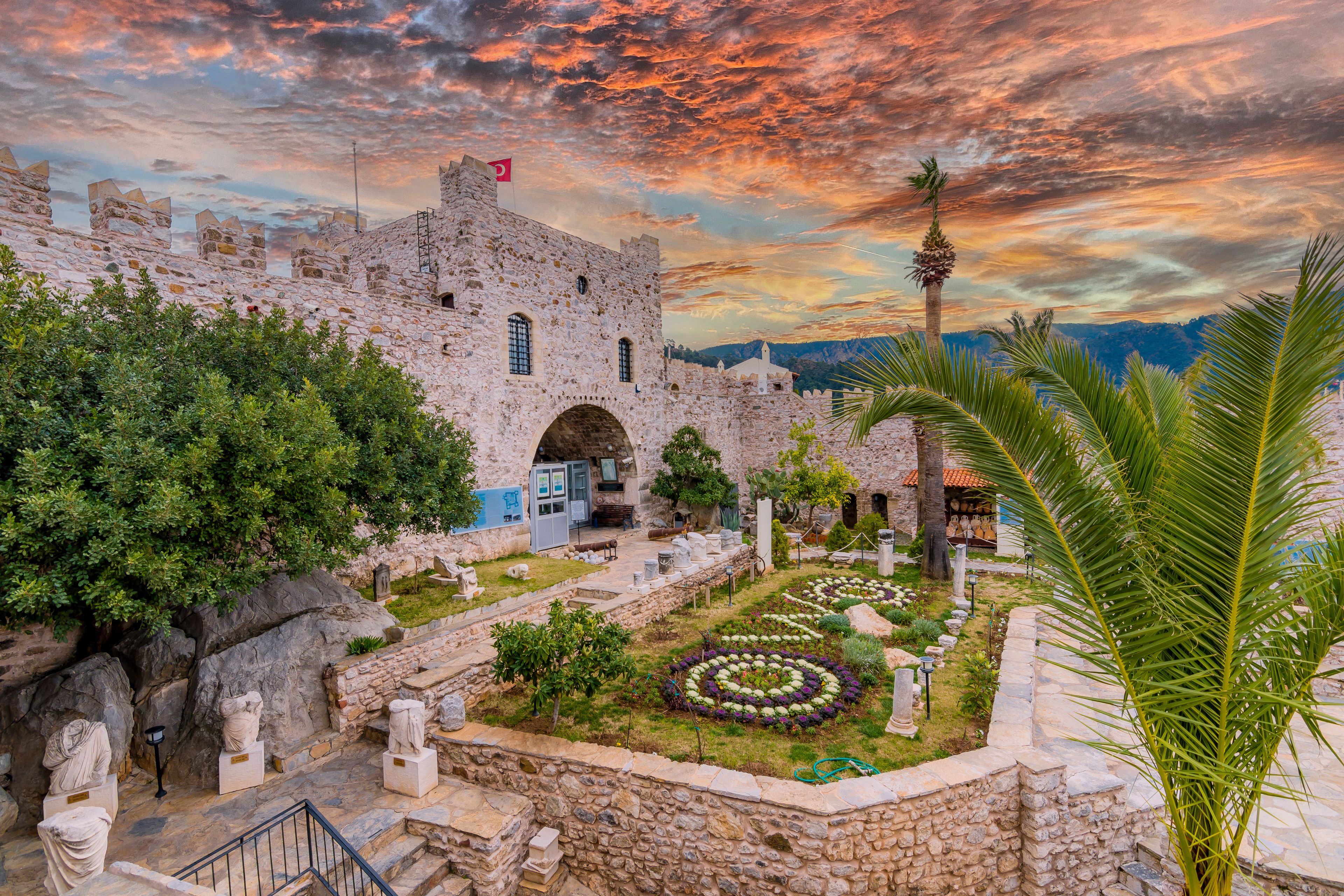 Un castillo de piedra histórico con una bandera, un jardín con estatuas y un cielo al atardecer.