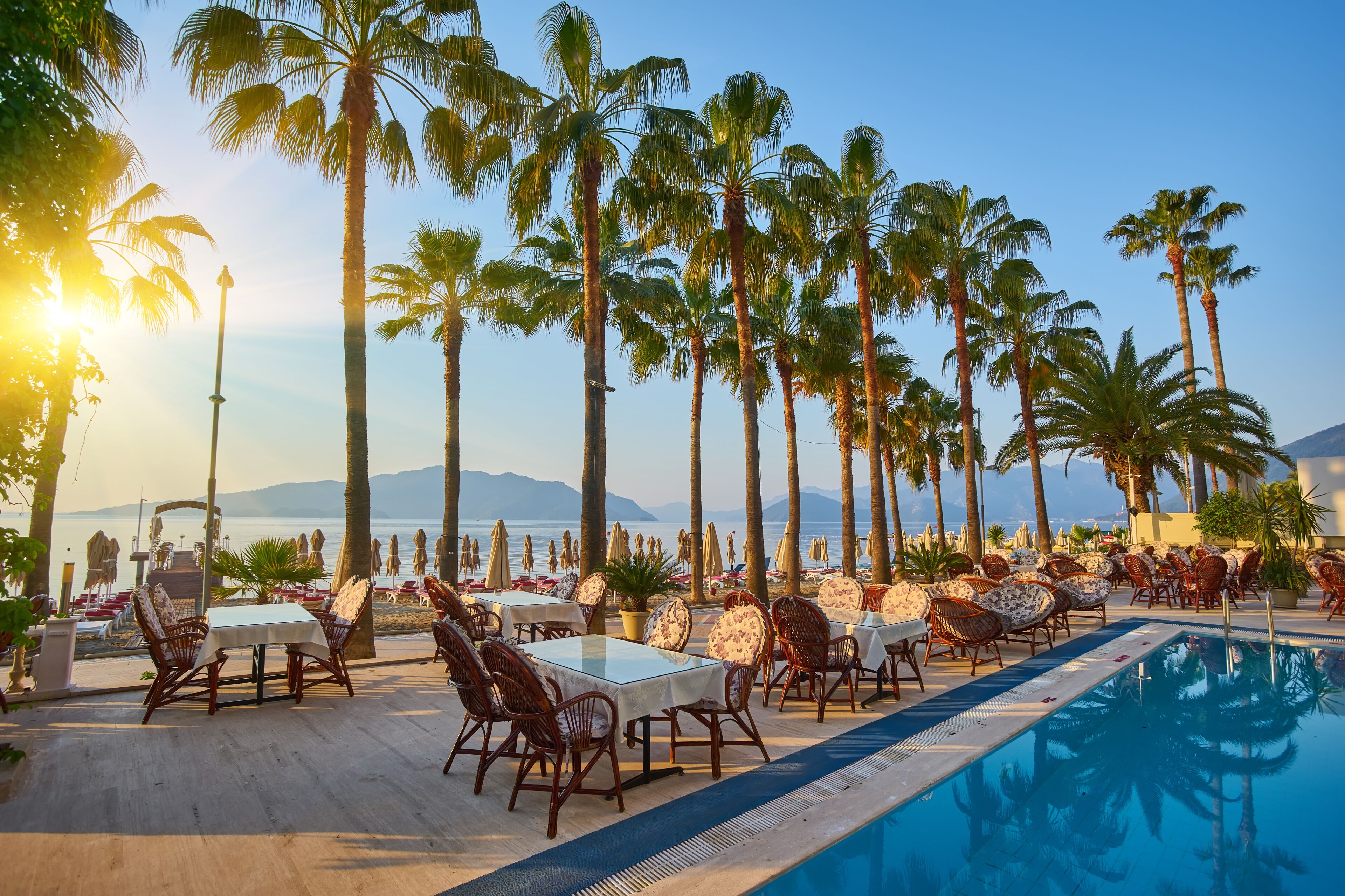 Zona junto a la piscina con mesas y sillas, palmeras y vista al mar.