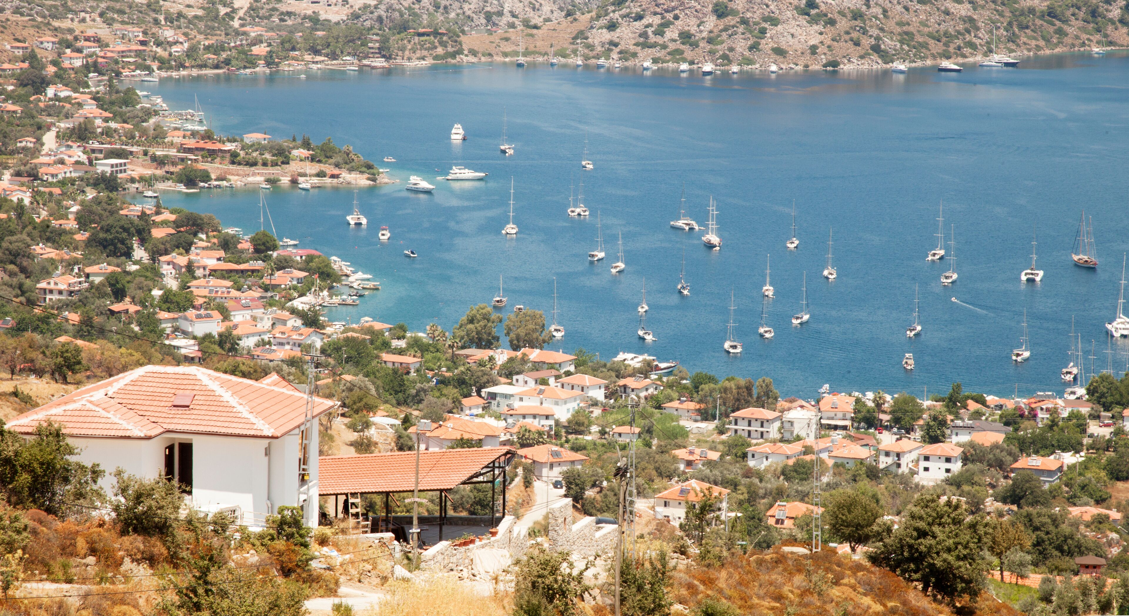 A coastal town with numerous sailboats anchored in the bay, surrounded by residential buildings and hilly terrain.