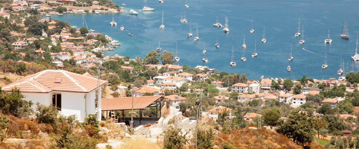 A coastal town with numerous sailboats anchored in the bay, surrounded by residential buildings and hilly terrain.