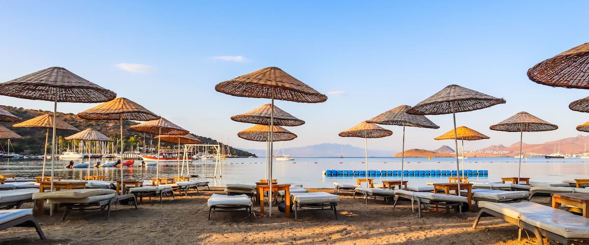 A beach with rows of sun loungers and thatched umbrellas, a calm sea, and boats in the background.