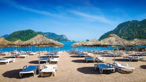 Tranquil beach setting in Marmaris with straw umbrellas, white sun loungers on sandy shore. Calm blue sea, picturesque mountains under clear sky. Welcome to Marmaris, Turkey concept image