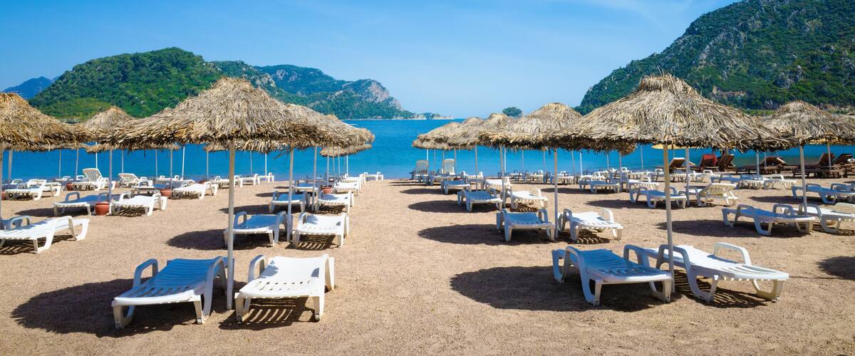 A beach with white lounge chairs and thatched umbrellas, surrounded by clear blue sea and green hills.