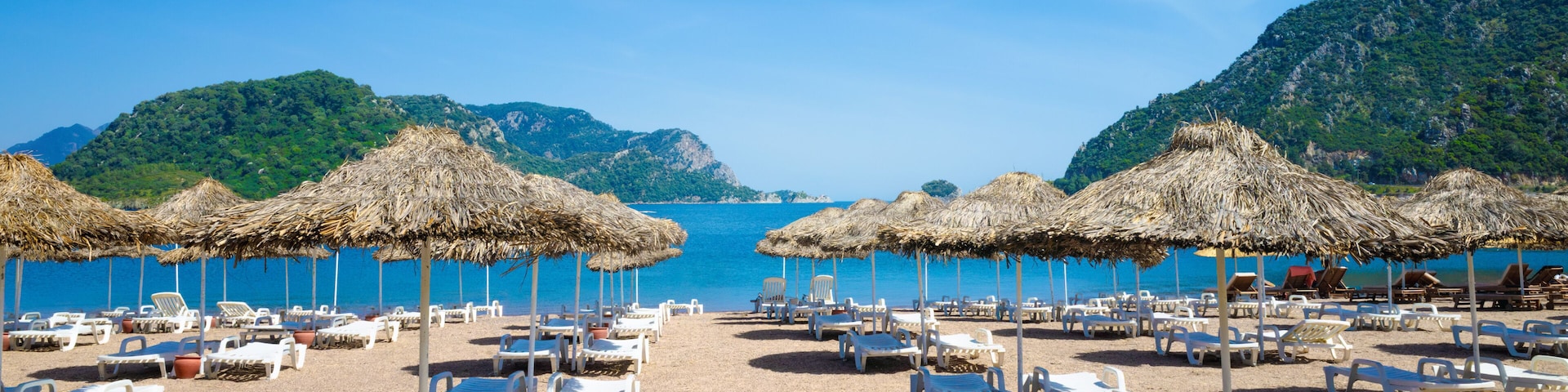 A beach with white lounge chairs and thatched umbrellas, surrounded by clear blue sea and green hills.