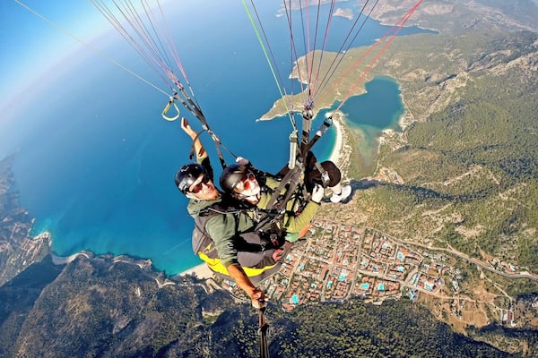 Zwei Personen gleiten mit einem Paragleiter über eine Küstenlandschaft, darunter eine Stadt.