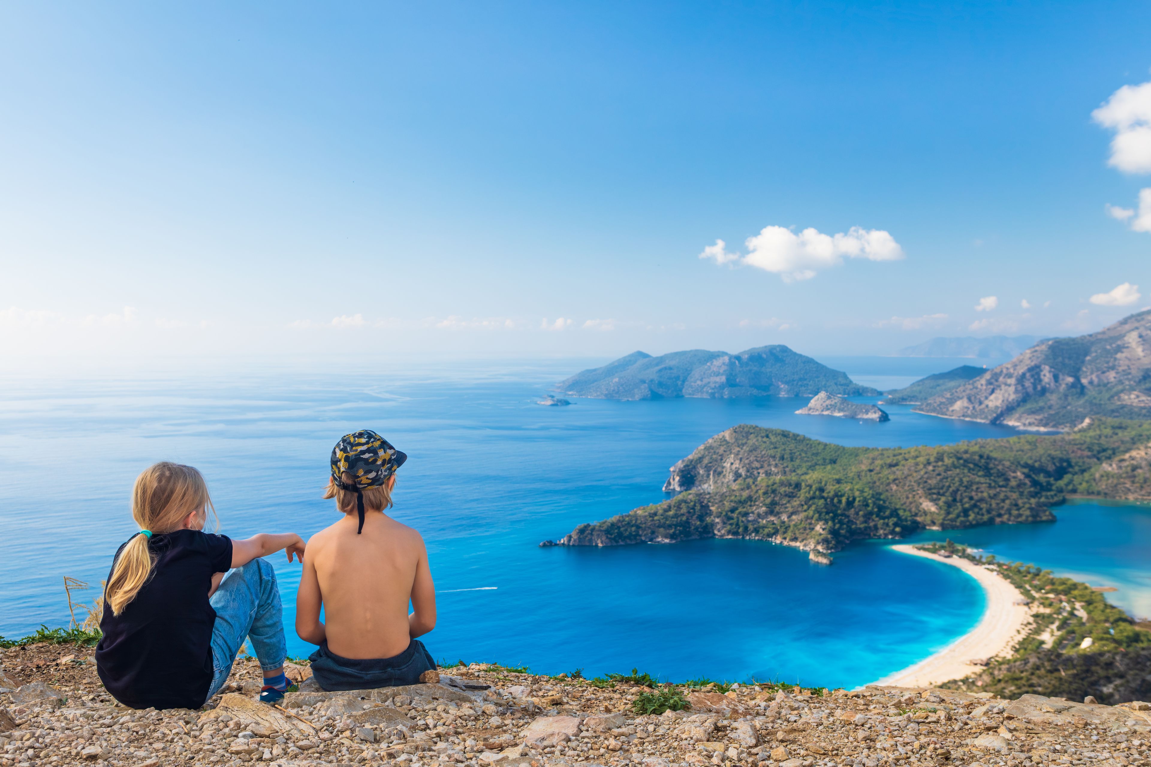 Two people sitting on a rocky outcrop overlooking a coastal landscape with clear blue waters and distant islands.
