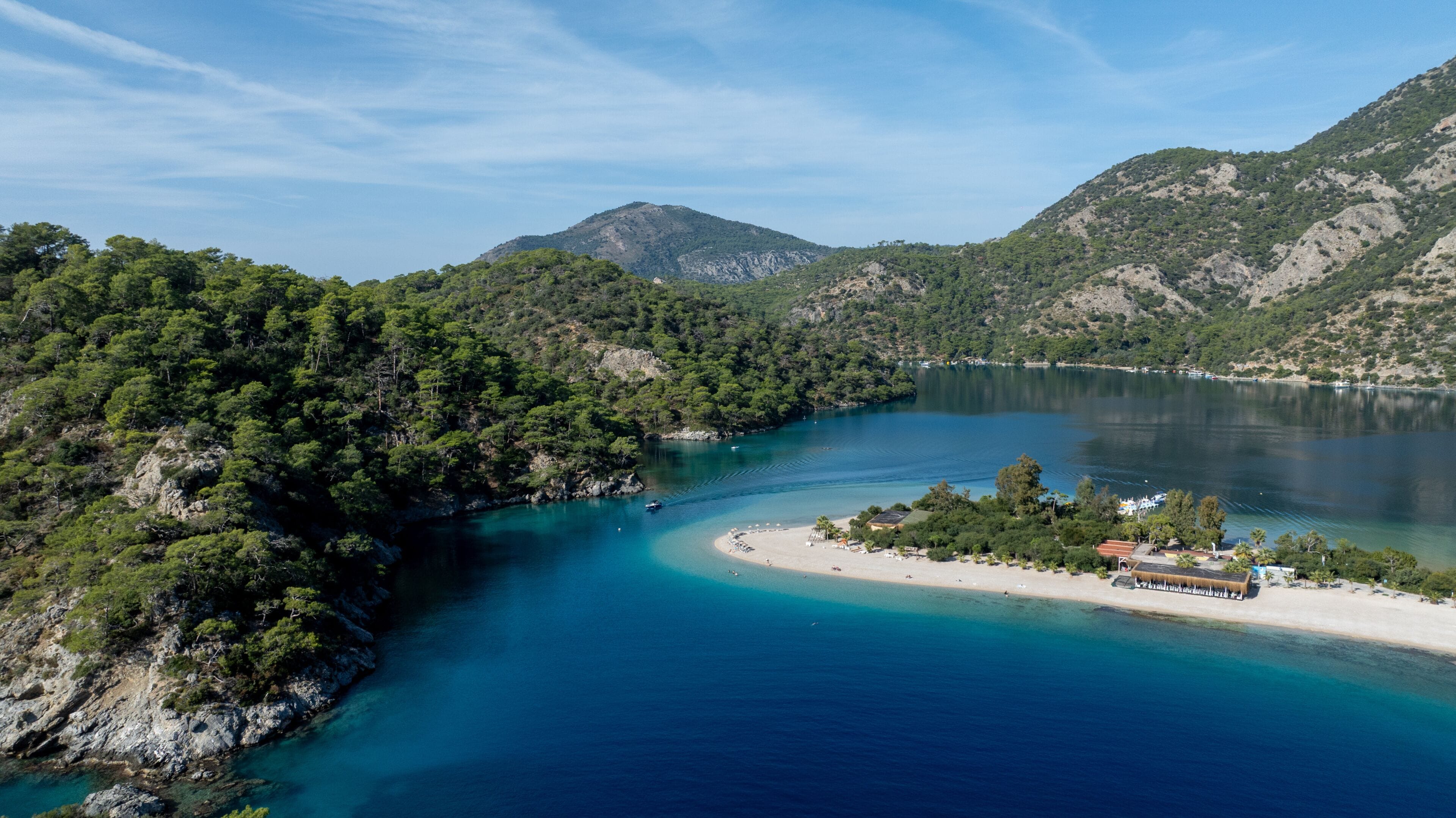 A coastal landscape with a clear blue sea, a sandy beach, and a mountainous backdrop.