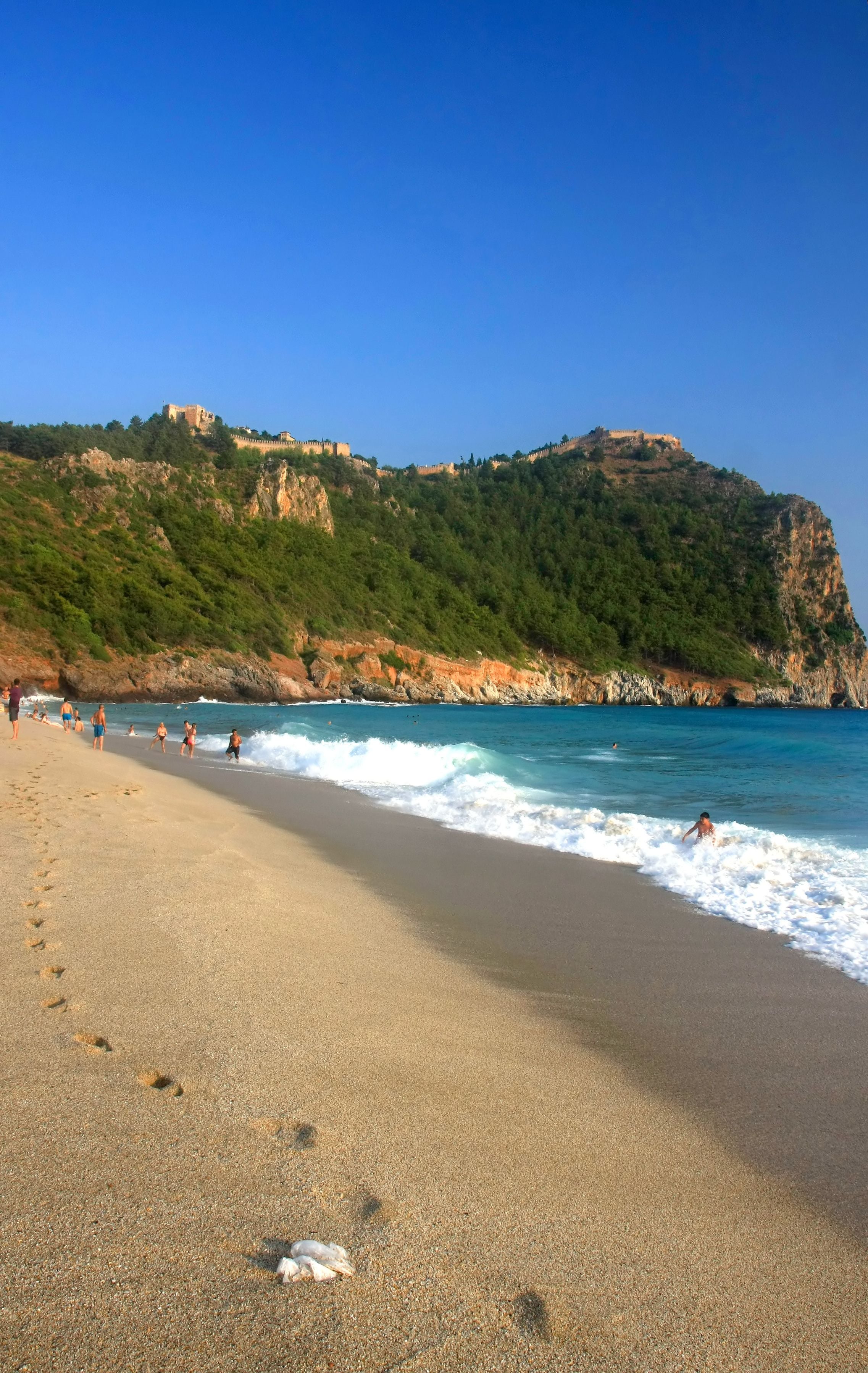 A beach with waves, footprints, and a castle on a hill.