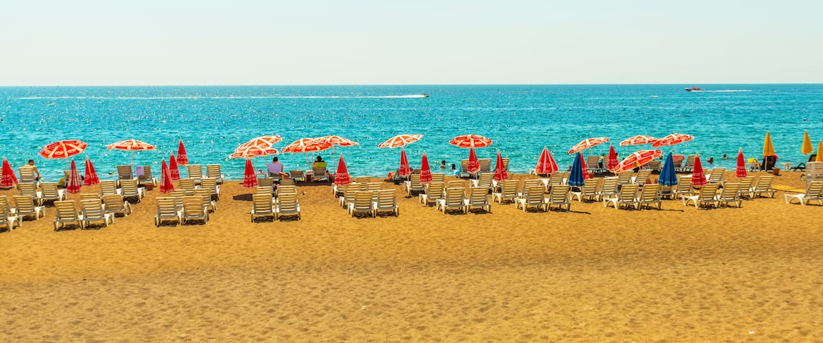 A beach with rows of sun loungers and umbrellas, a clear blue sea, and a boat in the distance.