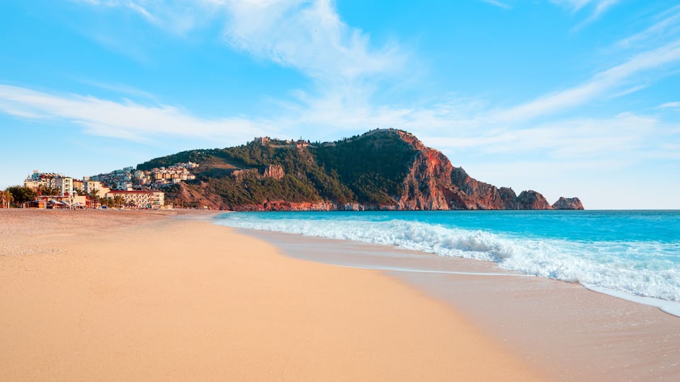 A beach with clear turquoise waters, a sandy shore, and a prominent rocky mountain in the background.