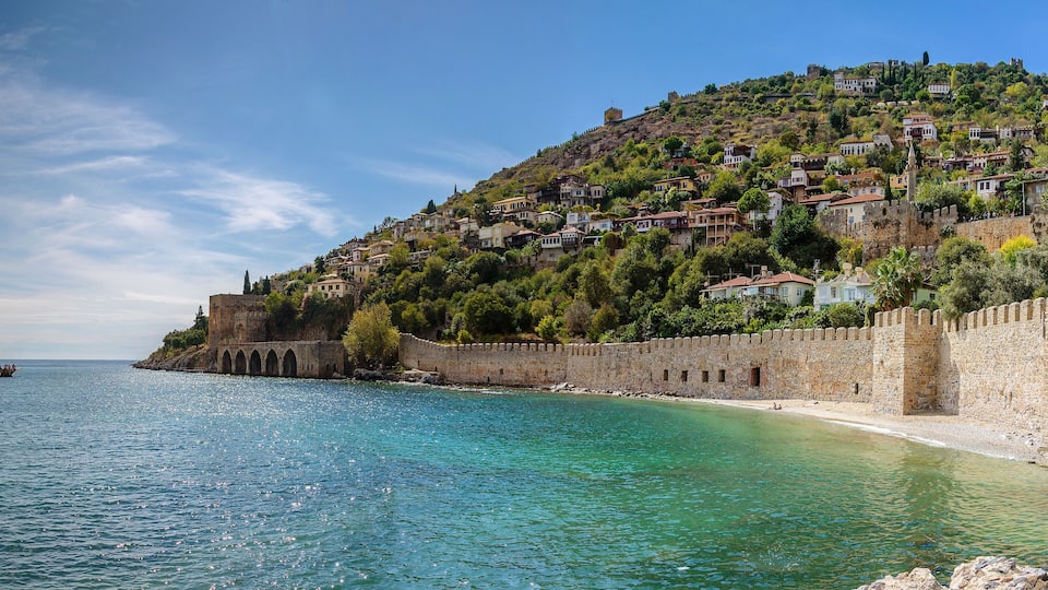 A coastal town with a historic stone wall, clear turquoise waters, and a boat on the sea.