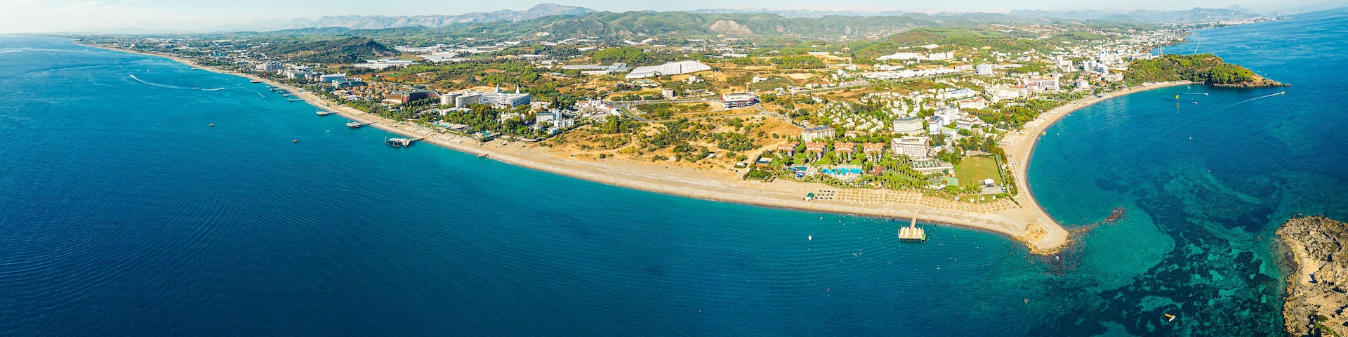 An aerial view of a coastal town with a beach, clear blue waters, and surrounding hills.