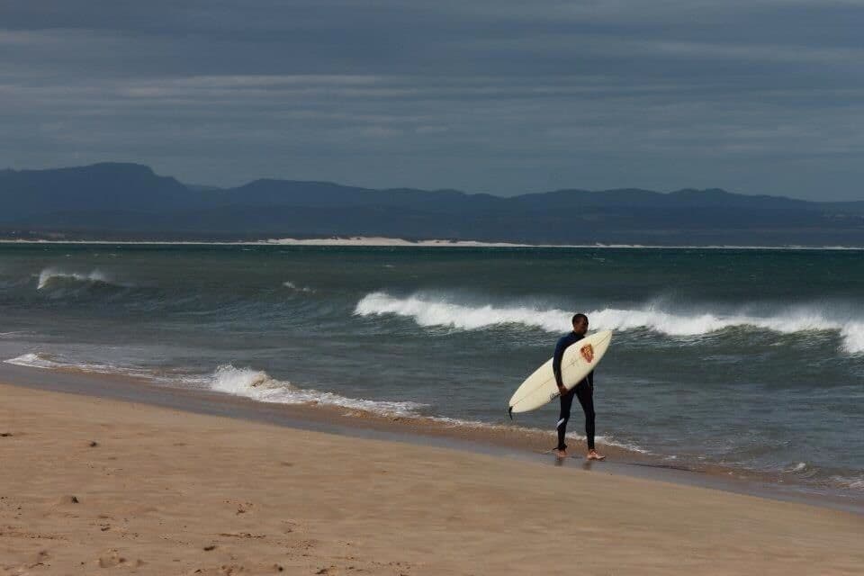 Una persona sostiene una tabla de surf en una playa, con olas y montañas al fondo.