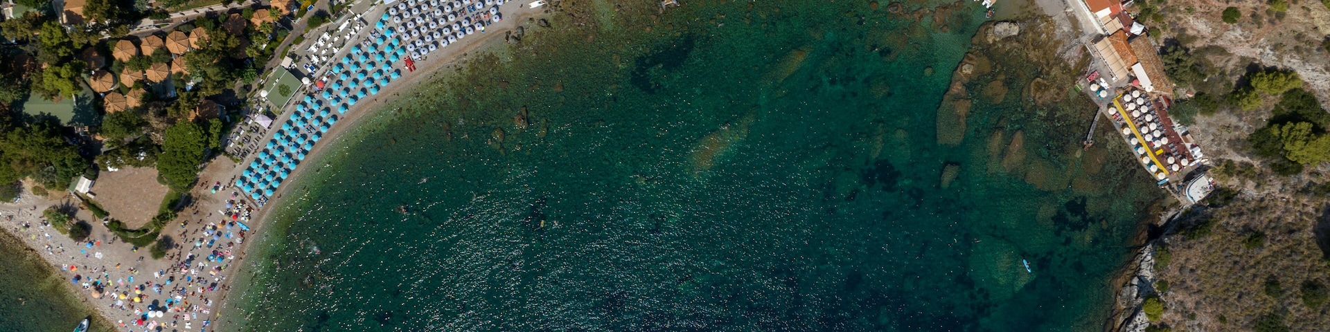 An aerial view of a coastal area with a beach, a swimming pool, and a densely packed row of umbrellas.
