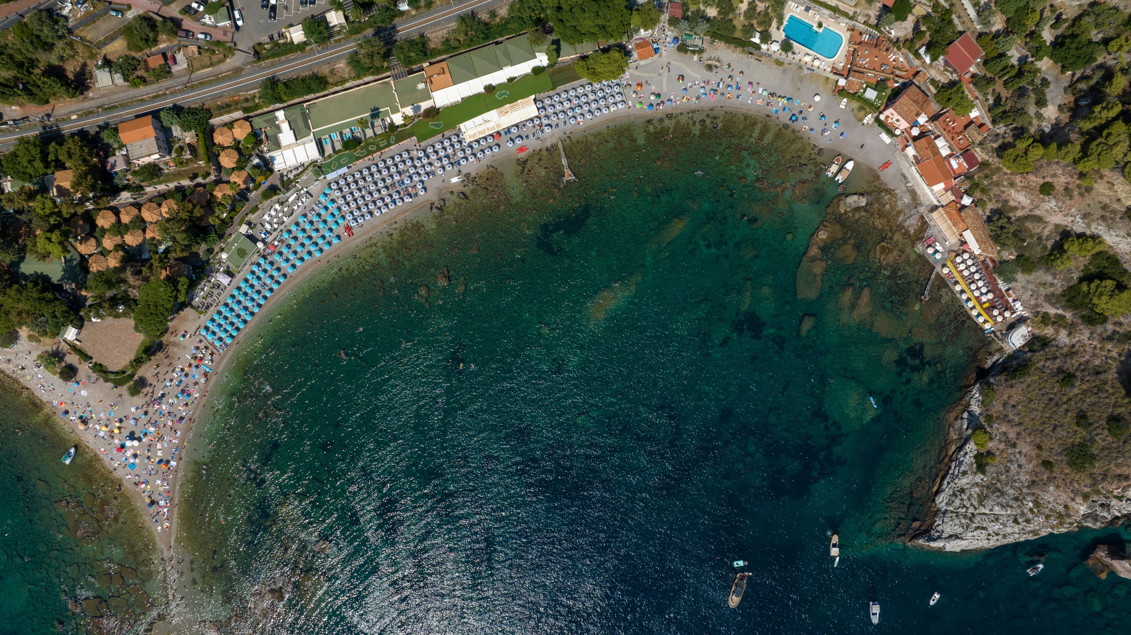 An aerial view of a coastal area with a beach, a swimming pool, and a densely packed row of umbrellas.